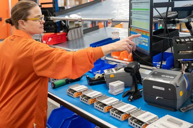 A woman in an orange smock assembling small orange industrial PCs (IPC) while touching a panel PC following a work instruction 